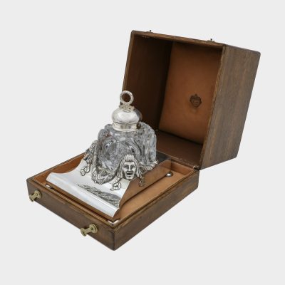 Faberge inkwell in original box, cut-glass inkwell on trapezoid form silver stand, front corners cast with theatrical masks