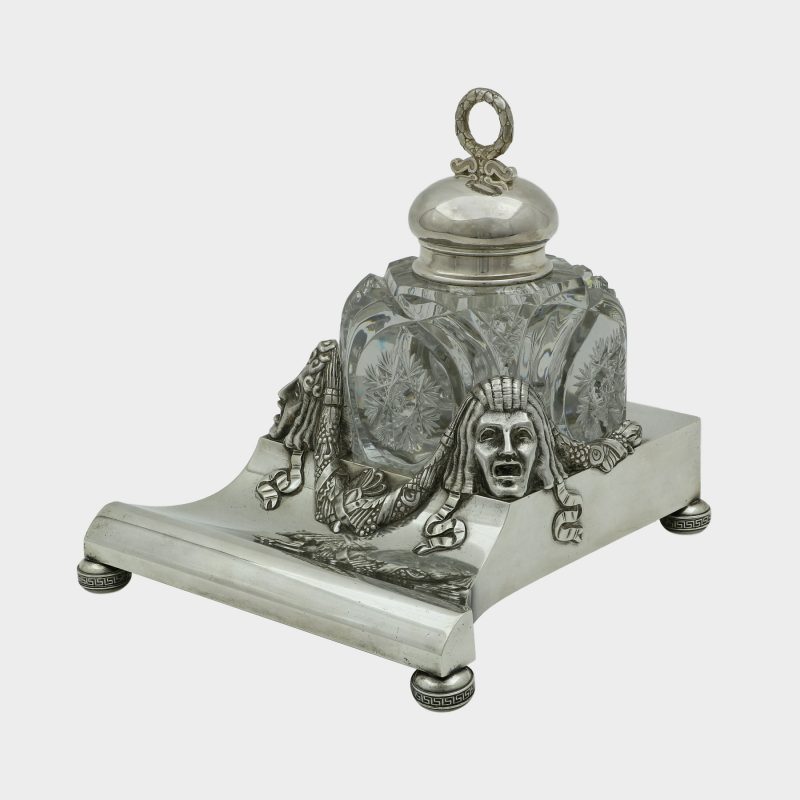 corner view on Faberge inkwell, cut-glass inkwell on trapezoid form silver stand, front corners cast with theatrical masks